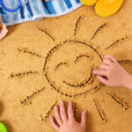 Child drawing a smiley sun in sand with towel, seashells and flip flops