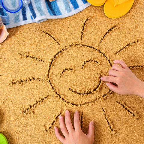 Child drawing a smiley sun in sand with towel, seashells and flip flops